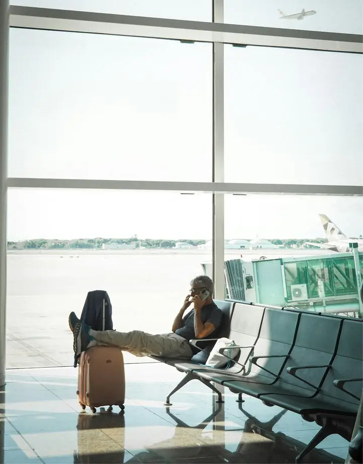 Man on the phone in an airport lounge, suitcase beside him, as a plane takes off